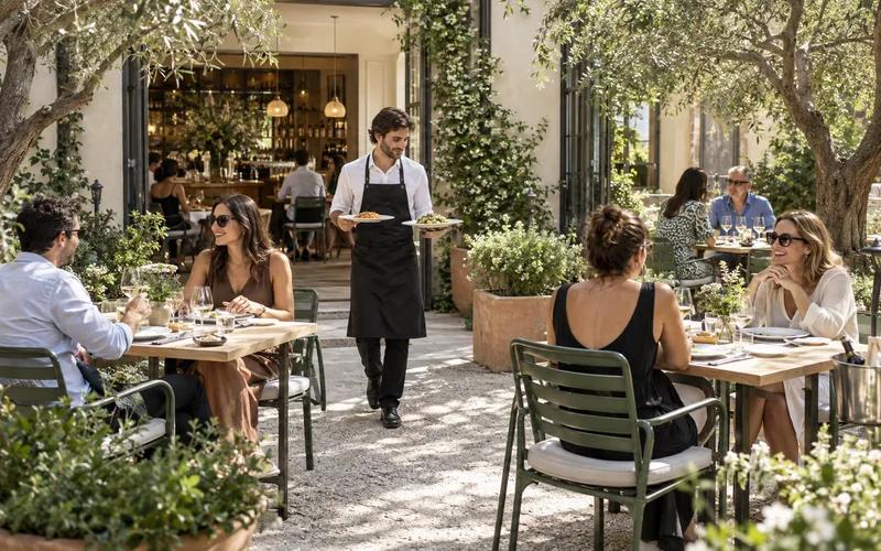 A server carrying dishes between dining tables on a leafy Italian restaurant patio.